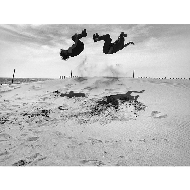 Two dancers jump in the air on the beach as if they are in midst of flipping